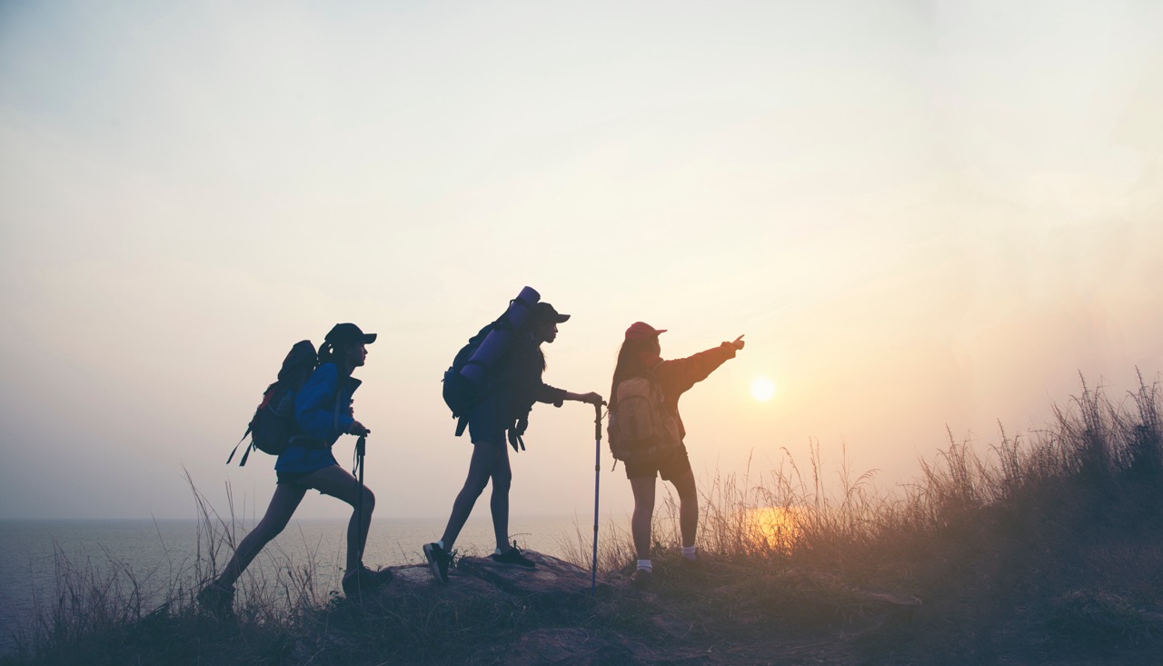 Silhouettes of three hikers with backpacks and trekking poles walking along a grassy ridge at sunset, with one hiker pointing toward the horizon.