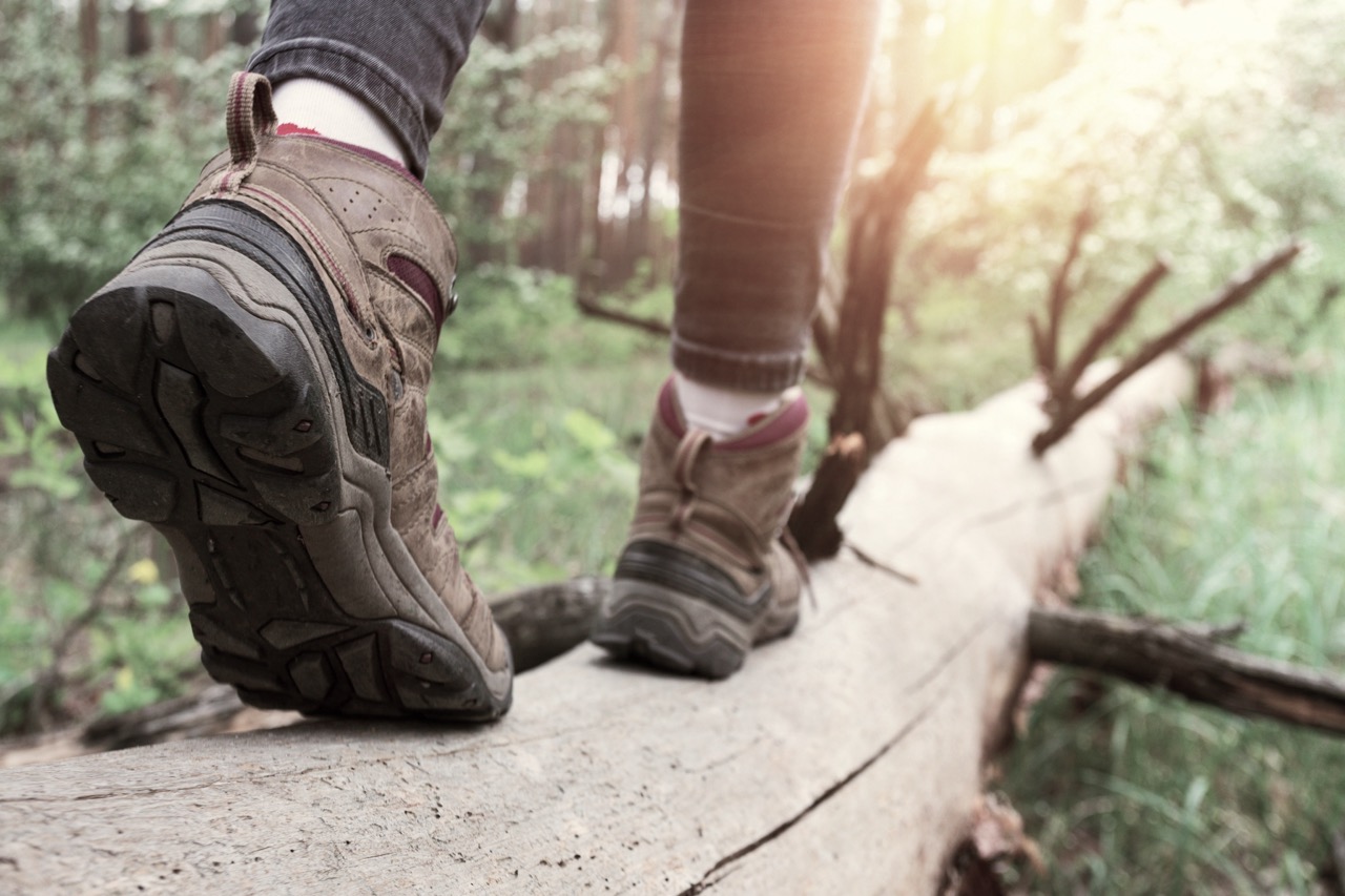Close-up of hiking boots walking along a fallen log in a forest, with blurred greenery and sunlight in the background — symbolizing balance, focus, and navigating the path ahead.