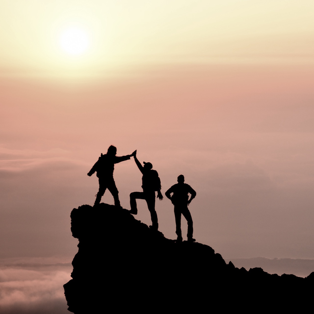 Three hikers standing on a rocky peak at sunset, one giving a high five while the others stand triumphantly against the sky.