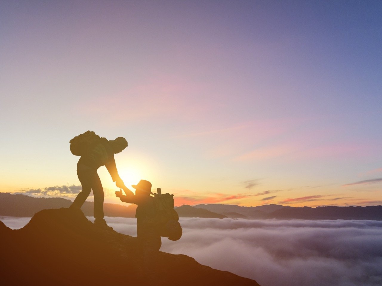 Silhouette of one hiker helping another up a mountain ridge at sunrise, with a glowing sky and clouds below.